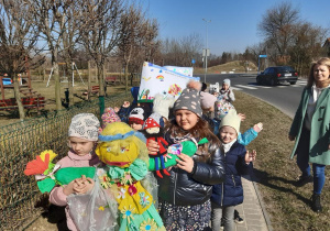 Dzieci maszerują ulicami miasta w przemarszu wiosennym, dwie dziewczynki trzyma w ręku kukłę Pani Wiosny oraz maskotkę Krasnala Hałabały.